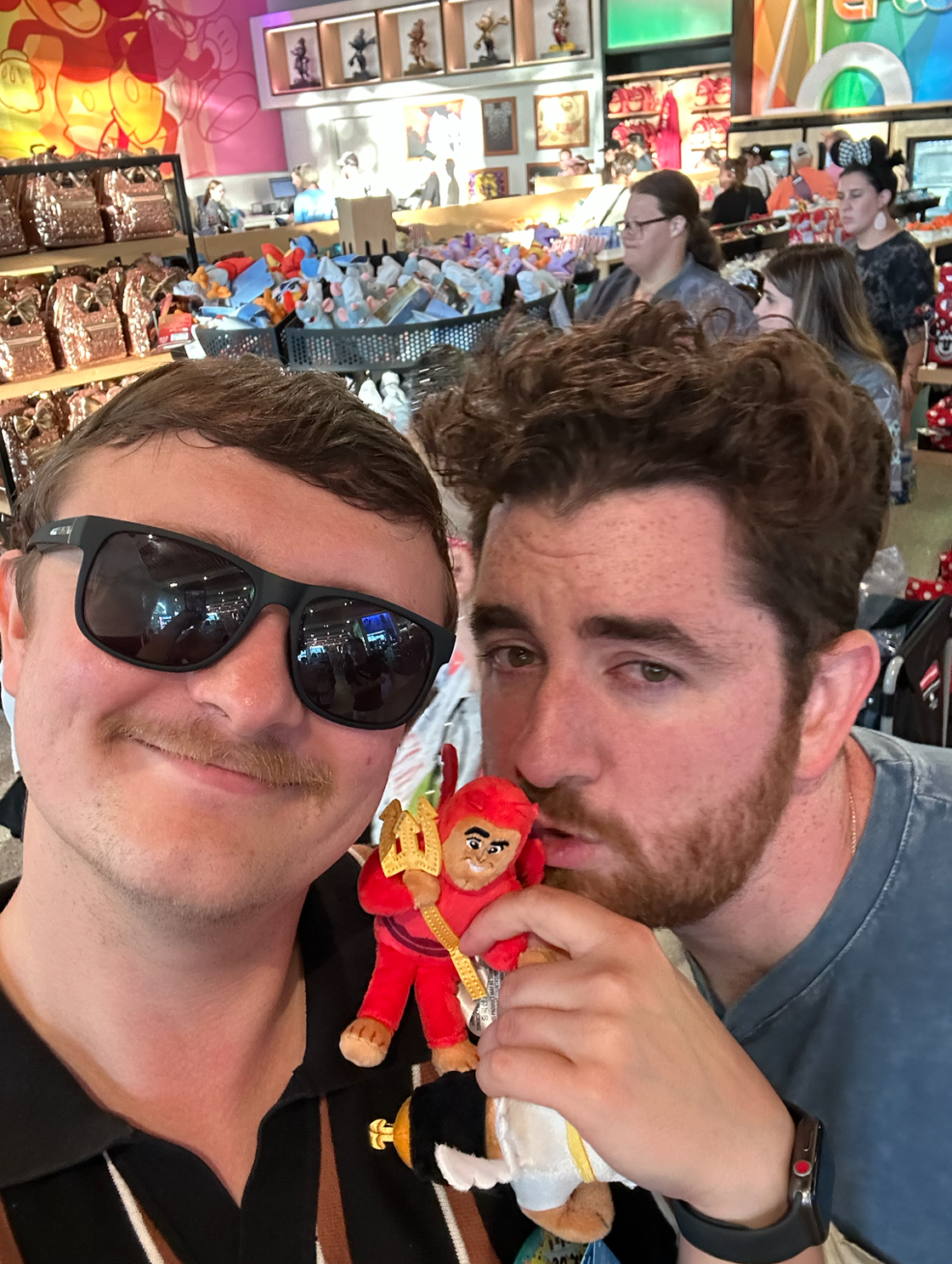 Two young men stand in the middle of a Disney store posing with a Kronk character plushie.
