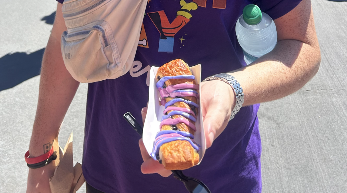 A man in a purple shirt stands holding the Cheshire Cat Tail pastry in a white container at Magic Kingdom in Disney World.