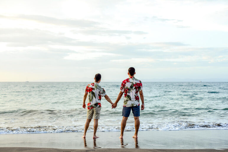 Two men stand on the beach looking out at the water in floral shirts.