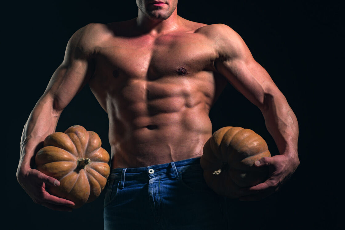 A muscular shirtless man poses in blue jeans and holds a pumpkin in each hand.