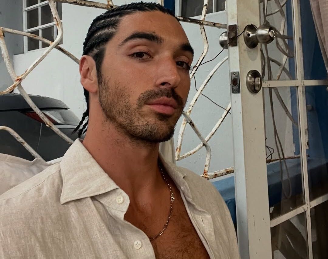 Taylor Zakhar Perez poses with his shirt open, wearing cornrows while visiting Puerto Rico, and smiling suggestively.