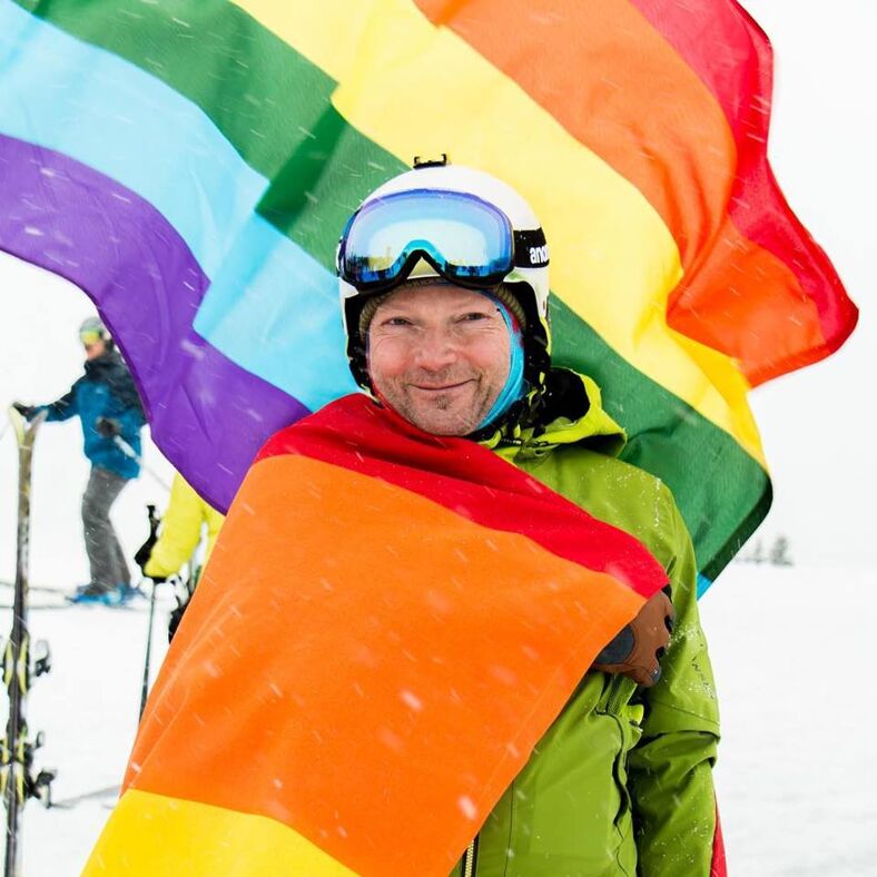 Man dressed in ski apparel in front of a rainbow pride flag. 