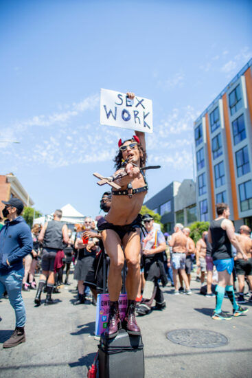 Person wearing a black leather harness stands on a speaker holding a crucifix and a sign that reads "sex work". 