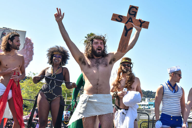 A man dressed as Jesus wearing a crown of thorns holding a crucifix that reads "say gay."