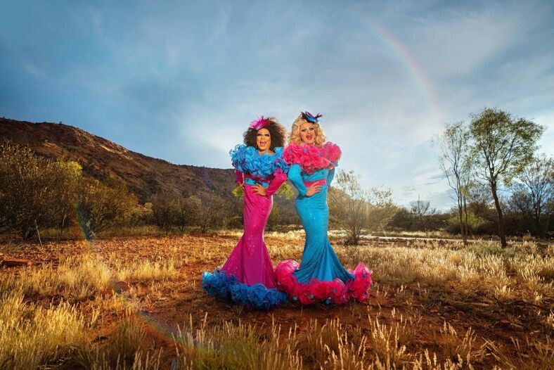 Two drag queens in pink and blue outfits standing back-to-back under a rainbow.